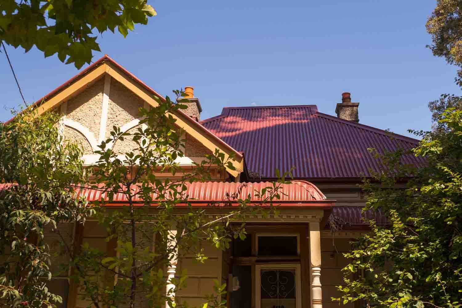 A Melbourne Victorian cottage with a red corrugated roof and brick chimneys, framed by garden trees